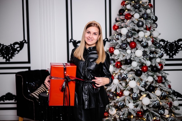 A beautiful woman stands by a garlanddecorated Christmas tree and opens a gift Next to it is a Christmas tree with boxes of gifts decorated New Year