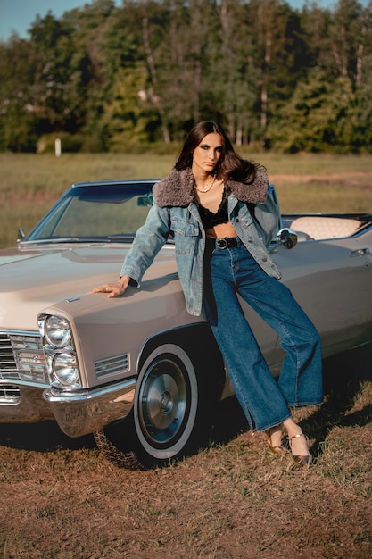 Foto una bella donna si trova accanto a una cabriolet in un campo vestita in jeans e una giacca con pelliccia bellissimo e elegante look