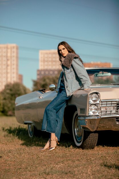 A beautiful woman stands next to a convertible in a field Dressed in jeans and a jacket with fur Beautiful and stylish look