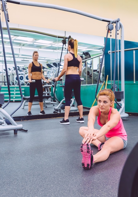 Beautiful woman stretching in a fitness center and female friend doing exercises with dumbbells