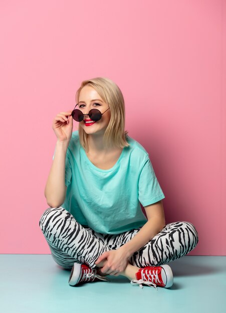 Beautiful woman in style clothes and sunglasses sitting on blue floor near pink wall