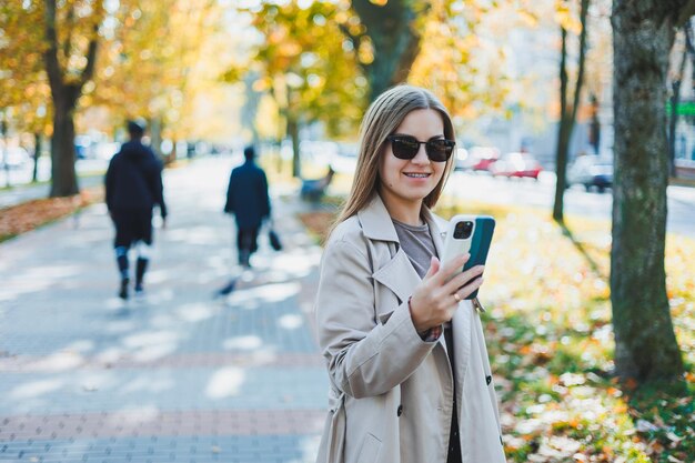 A beautiful woman in sunglasses is walking in the autumn park and talking on the phone Joyful girl on the autumn alley in the city