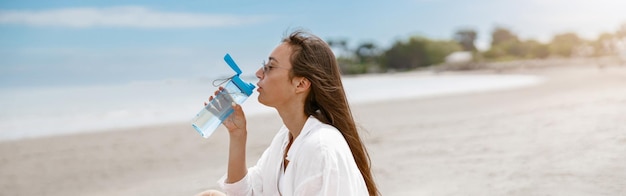 サングラスをかけた美しい女性が海の近くに座って水を飲み、休暇中にリラックス