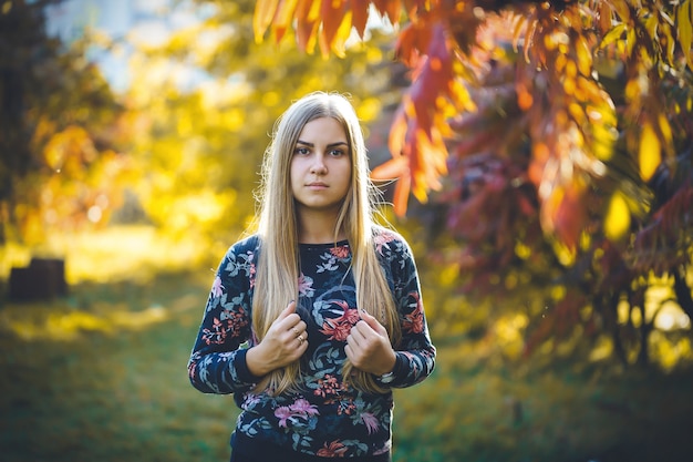 落ち葉と秋の自然の中で座っているセーターとズボンの美しい女性。明るい若い長い髪の少女は、秋に赤黄色の葉で公園でリラックスします。ロマンチックなコンセプト。