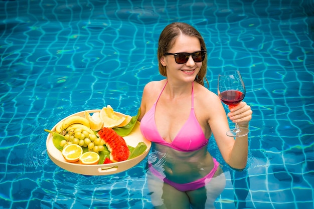 Photo beautiful woman in the swimming pool with glass of wine and floating tray of fruits