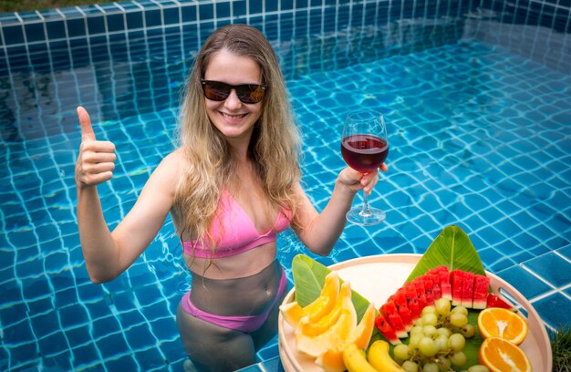ワインのグラスとフルーツの浮かぶトレイを持つプールにいる美しい女性