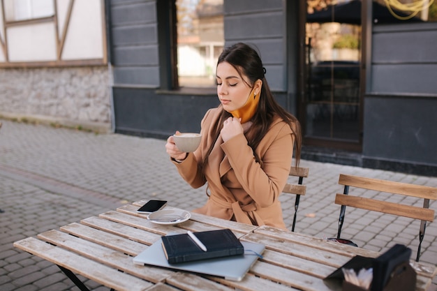 Bella donna si toglie la maschera protettiva per bere il caffè. donna seduta all'aperto sulla terrazza e bere un caffè.