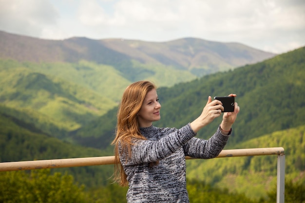 緑の山々を背景にスマートフォンで写真を撮る美しい女性