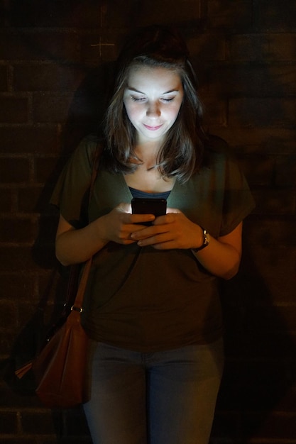 Photo beautiful woman text messaging against brick wall at night
