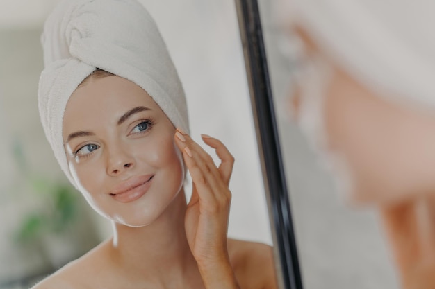 Beautiful woman touches face gently looks at her reflection in mirror enjoys softness of skin stands with wrapped bath towel on head has minimal makeup and manicure Beauty treatment concept