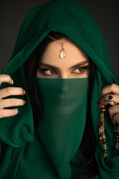 Beautiful woman in traditional costume. Stylish portrait of young girl with perfect bright makeup wearing traditional jewelry standing indoors against dark background. The model looking away