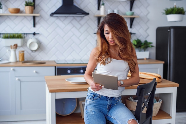 Beautiful woman uses tablet computer while sitting on stylish bright kitchen.