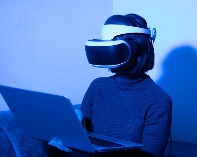 Beautiful woman using headset typing on laptop at home in blue light. The concept of the virtual universe. Freelancer in the virtual space. Business success concept