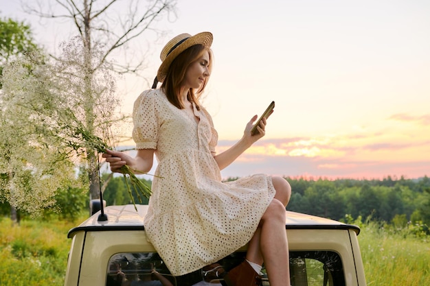 車の夏の風景の背景の外に座ってスマート フォンを使用して美しい女性