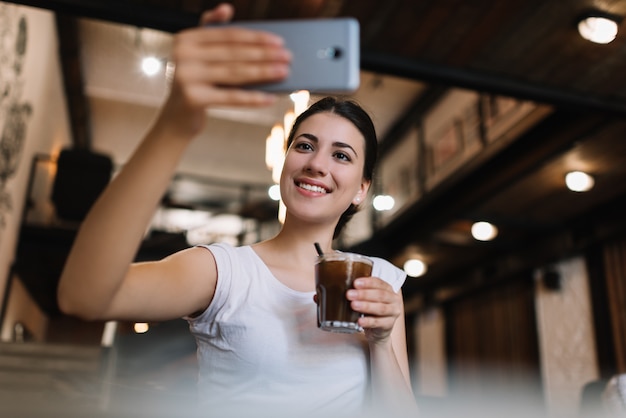 スマートフォンを使用して、selfieを取って、カフェでカクテルを飲んで美しい女性。ブロガーストリーミングビデオをオンラインで笑顔