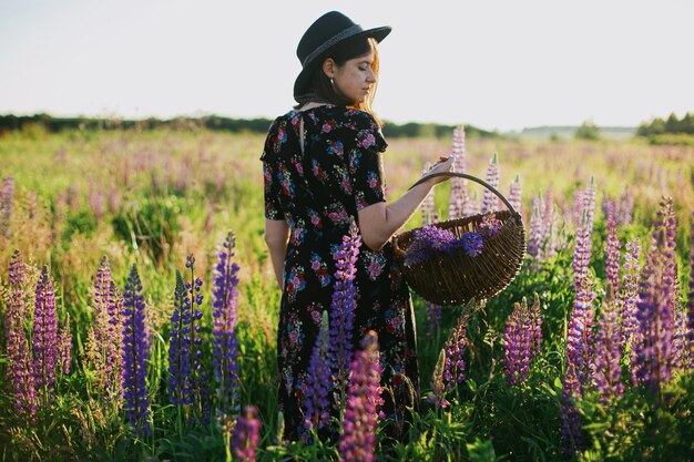 Photo beautiful woman walking in sunny lupine field with rustic basket and flowers tranquil moment