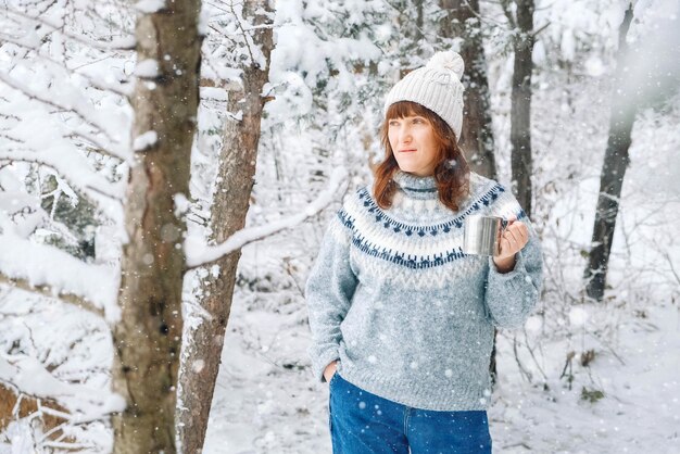 Beautiful woman in a warm hat holding cup of tea in her hands and standing in snowcovered forest