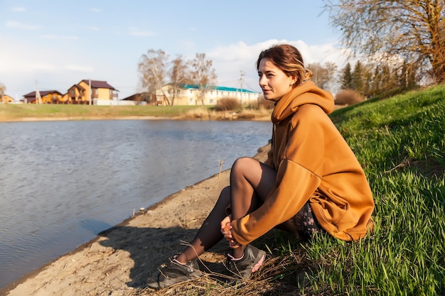 A beautiful woman in a warm sweatshirt dress and sneakers is sitting on the shore of the lake resting enjoying nature looking at the water on a warm summer day