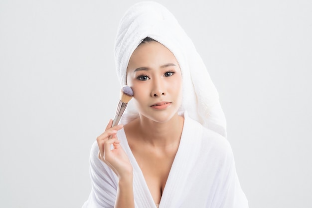 Beautiful woman wearing bathrobe with towel with towel on head is using a makeup brush makeup her after bathingon white background.