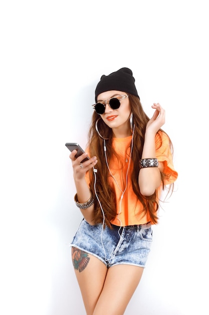 Beautiful woman wearing in black hat and white tshirt listening music near white wall holding a cell