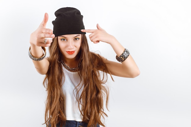 Beautiful woman wearing in black hat and white tshirt standing near white wall shows the gun fingers