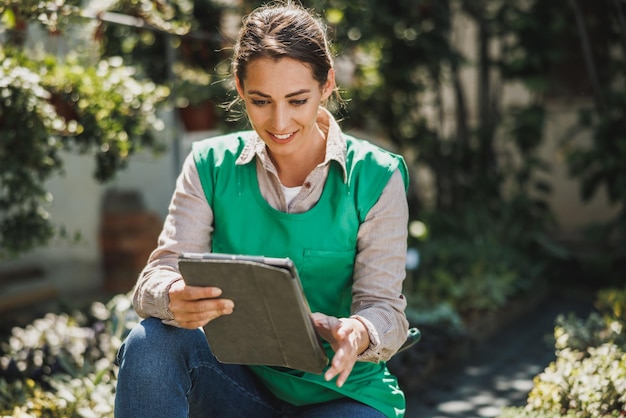 Beautiful woman wearing a green apron holding digital tablet and checking flowers in a garden center. Woman entrepreneur.