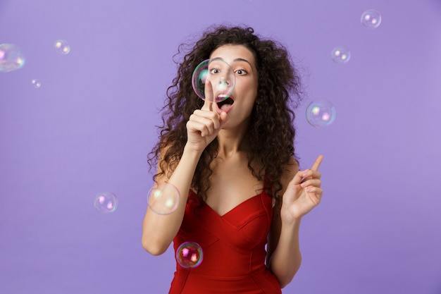 beautiful woman wearing red dress laughing and standing under falling soap bubbles, isolated over violet wall