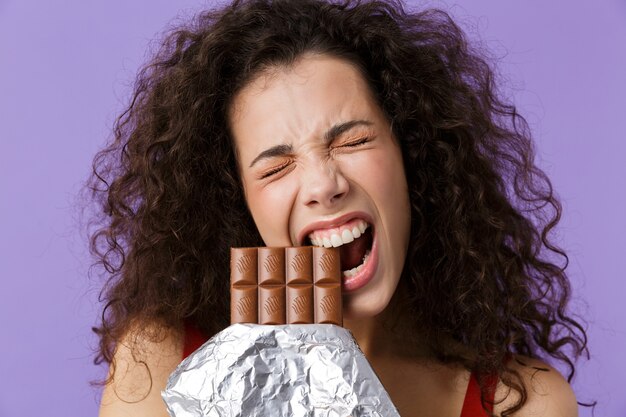 beautiful woman wearing red dress smiling and eating chocolate, standing isolated over violet wall