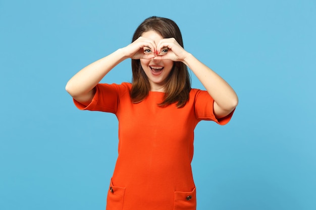 Beautiful woman wearing red orange dress holding hands near eyes, imitating glasses or binoculars isolated over blue background, studio portrait. People lifestyle fashion concept. Mock up copy space.