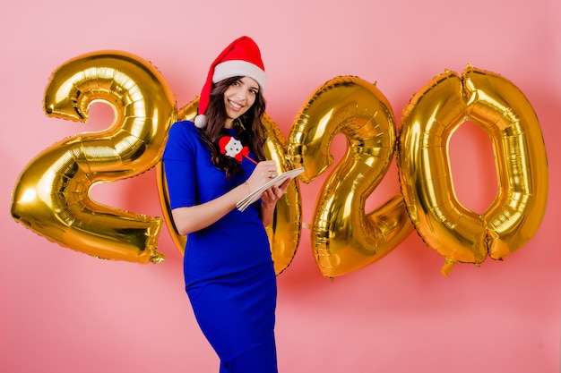 Beautiful woman wearing santa hat writing plans in notebook front of new year 2020 balloons isolated over pink