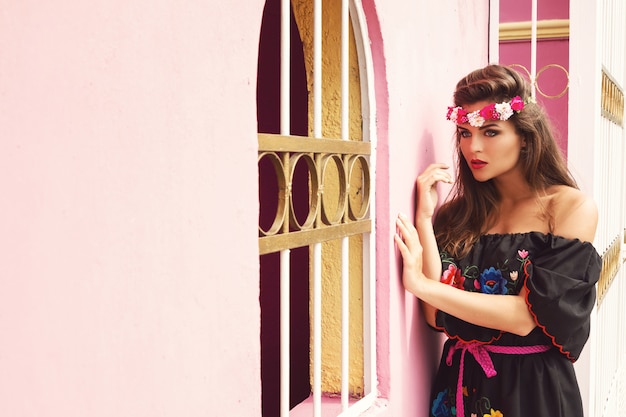 伝統的なメキシコのドレスを着ている美しい女性がピンクの壁の横にポーズをとってください。