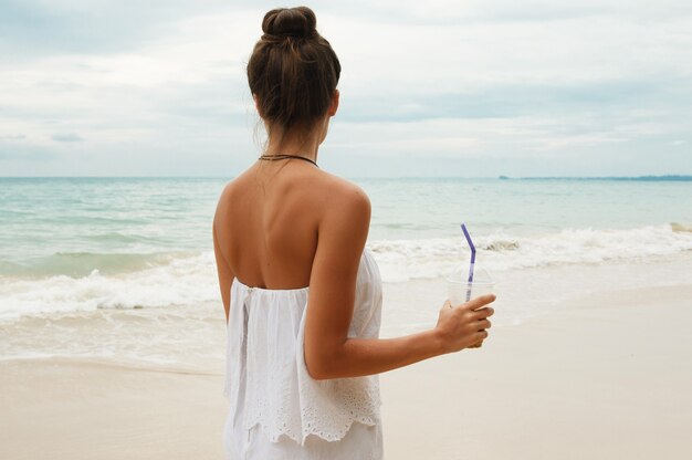 Bella donna che indossa tuta bianca a piedi sulla spiaggia con un bicchiere di caffè ghiacciato