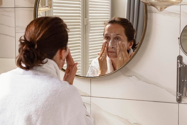 Красивая женщина в белом халате моет кожу лица в ванной с помощью зеркалаУход за кожей