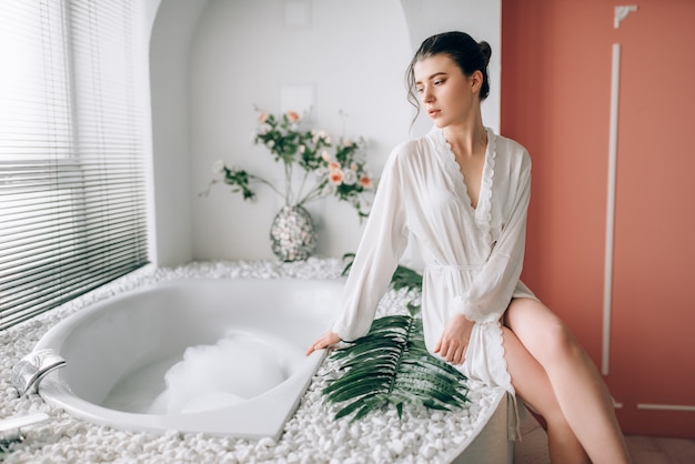 Beautiful woman in white bathrobe sitting on the edge of the bath with foam. Bathroom interior with window