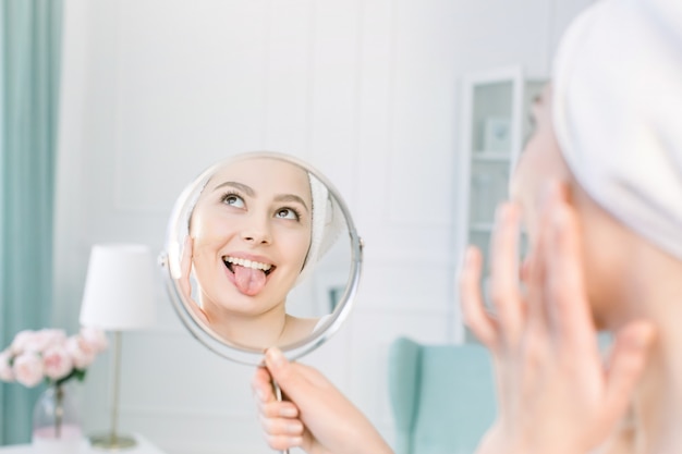 Красивая женщина в белом халате и полотенце смотрит на свою идеальную кожу в зеркале, показывает язык и наносит тональную крем-основу на лицо