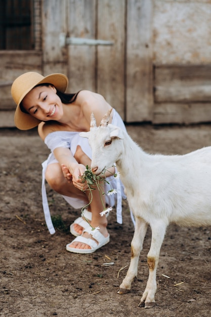 Bella donna e vestito bianco e con un cappello beige tra le capre in una fattoria ecologica