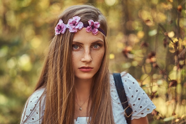 A beautiful woman in a white dress and purple wreath on head posing in a green autumn forest.