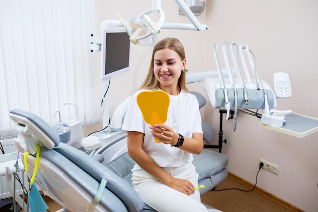 白いTシャツを着た美しい女性が歯科医院に座って鏡を見て、微笑んで歯ブラシを手に持っています。中かっこを持つ女の子は、口腔ケアを示しています。歯科、歯科治療