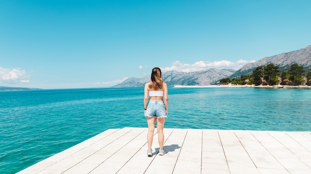 Bella donna in argomento bianco e pantaloncini di jeans da solo in riva al mare, guardando verso l'oceano blu