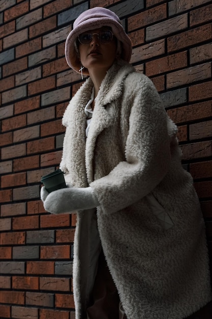 Bella donna con cappello d'inverno e cappotto di pelliccia sullo sfondo di un muro di mattoni per le strade della città in inverno
