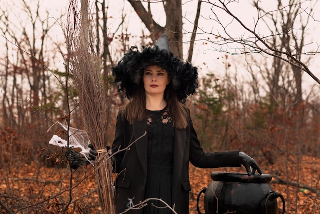 Beautiful woman in witches hat and costume holding broom near the cauldron in autumn forest Halloween concept Selective focus