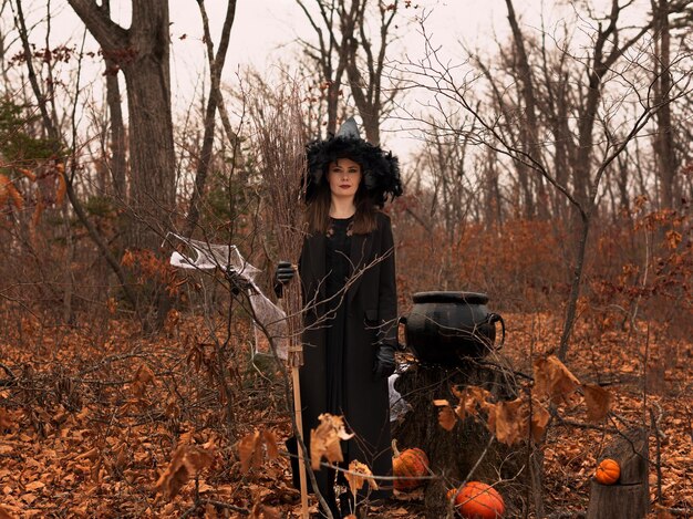 Foto bella donna in cappello da strega e costume che tiene scopa vicino al calderone nella foresta autunnale concetto di halloween fuoco selettivo