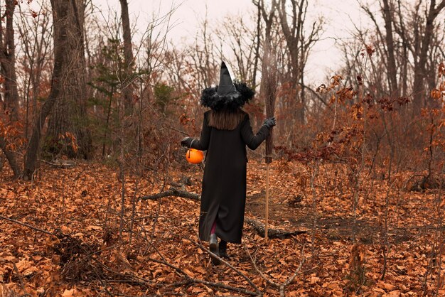 Beautiful woman in witches hat and costume holding orange plastic pumpkin and broom in autumn forest Halloween concept Selective focus