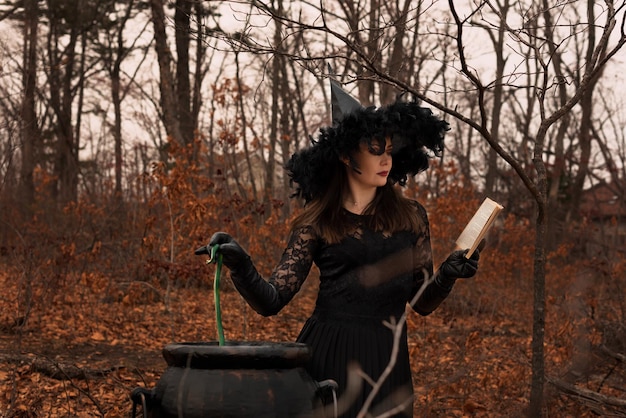 Beautiful woman in witches hat and costume preparing a potion in cauldron in autumn forest Halloween concept Selective focus