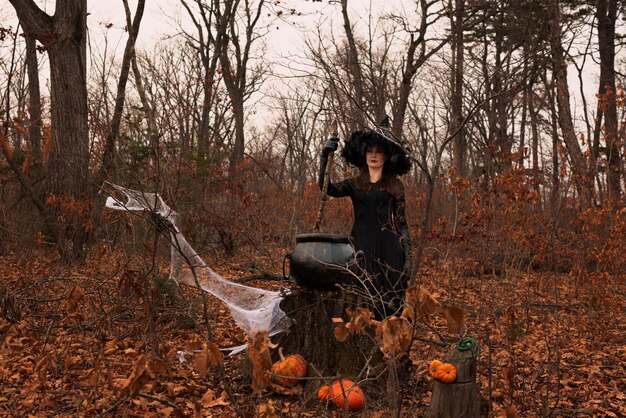 Красивая женщина в шляпе ведьмы и костюме готовит зелье в котле в осеннем лесу Концепция Хэллоуина Выборочный фокус