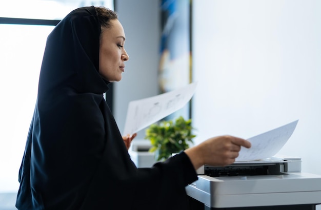 Beautiful woman with abaya dress working and printing documents. Middle aged female employee at work in a business office in Dubai. Concept about middle eastern cultures and lifestyle