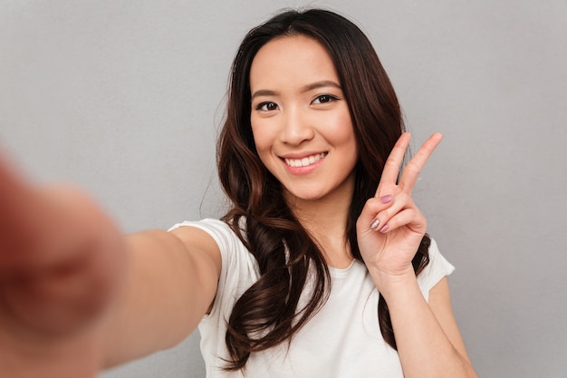 Красивая женщина с азиатской внешностью, принимая селфи и показывая знак победы с идеальной улыбкой, изолированных на серую стену