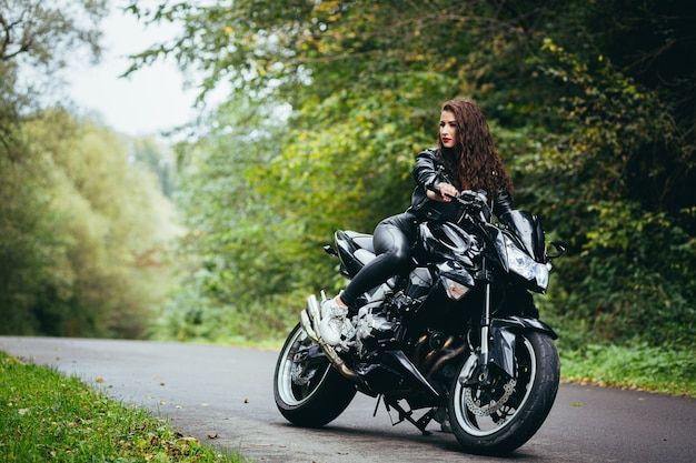 Красивая женщина, с черными вьющимися волосами, в черной кожаной куртке, сидит на спортивном мотоцикле