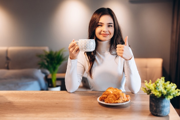 Bella donna con capelli neri lucidi che beve caffè durante la colazione. ritratto dell'interno della ragazza sveglia del brunette che mangia croissant e che gode del tè nella mattina.