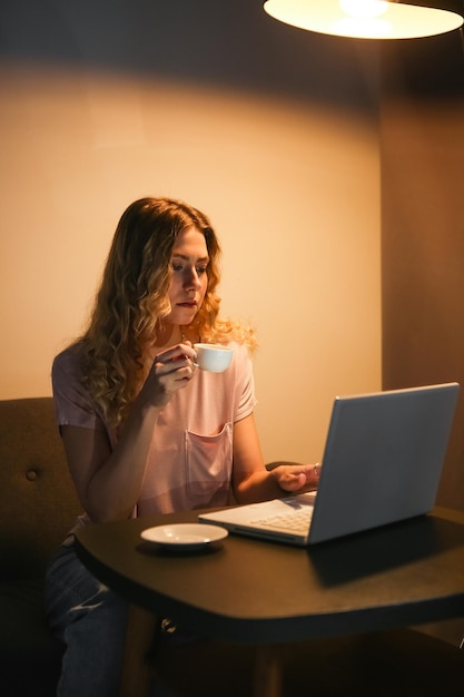 夕方にラップトップで作業しているコーヒーのカップを持つ美しい女性暗い部屋でノートブックを持つ女の子ディ